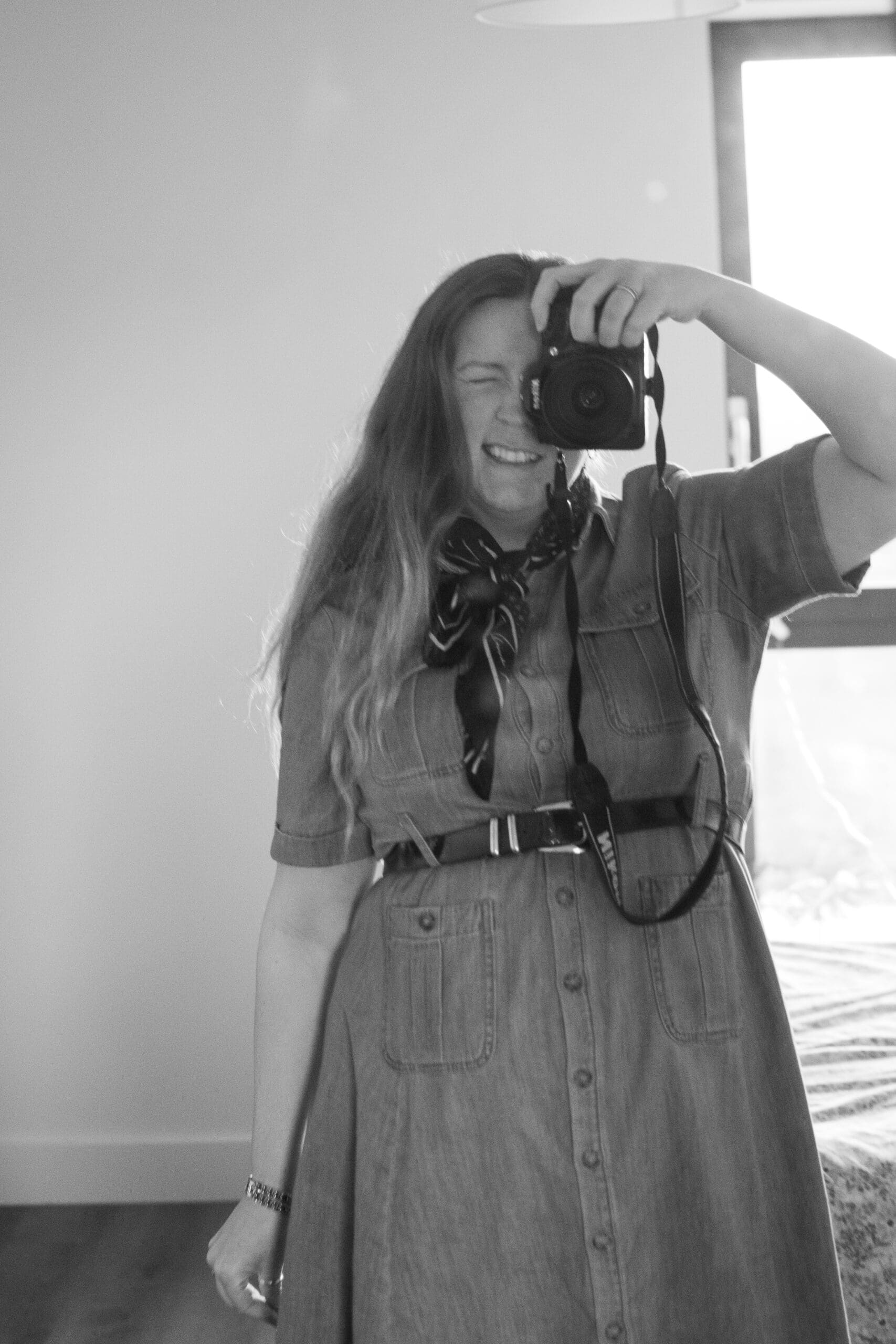 Andi holds up a camera to take a photograph. She is wearign a denim dress with her hair down, a belt and a silk scarf tied around her neck.