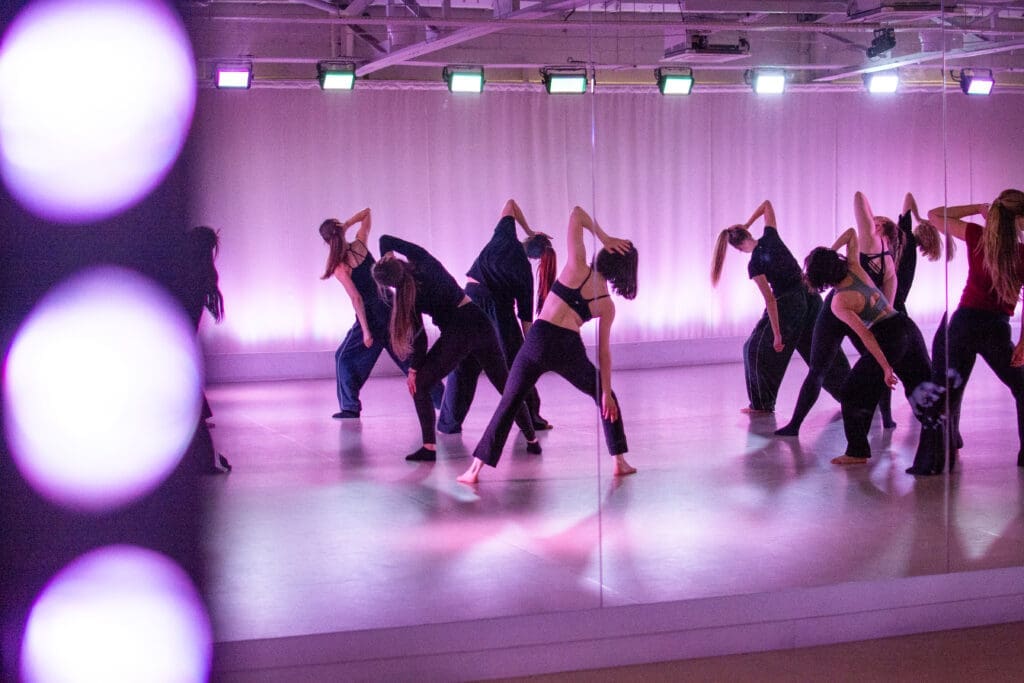 Dancers in a studio filled with purple light are leaning alternate directions with thier arms up in the air.
